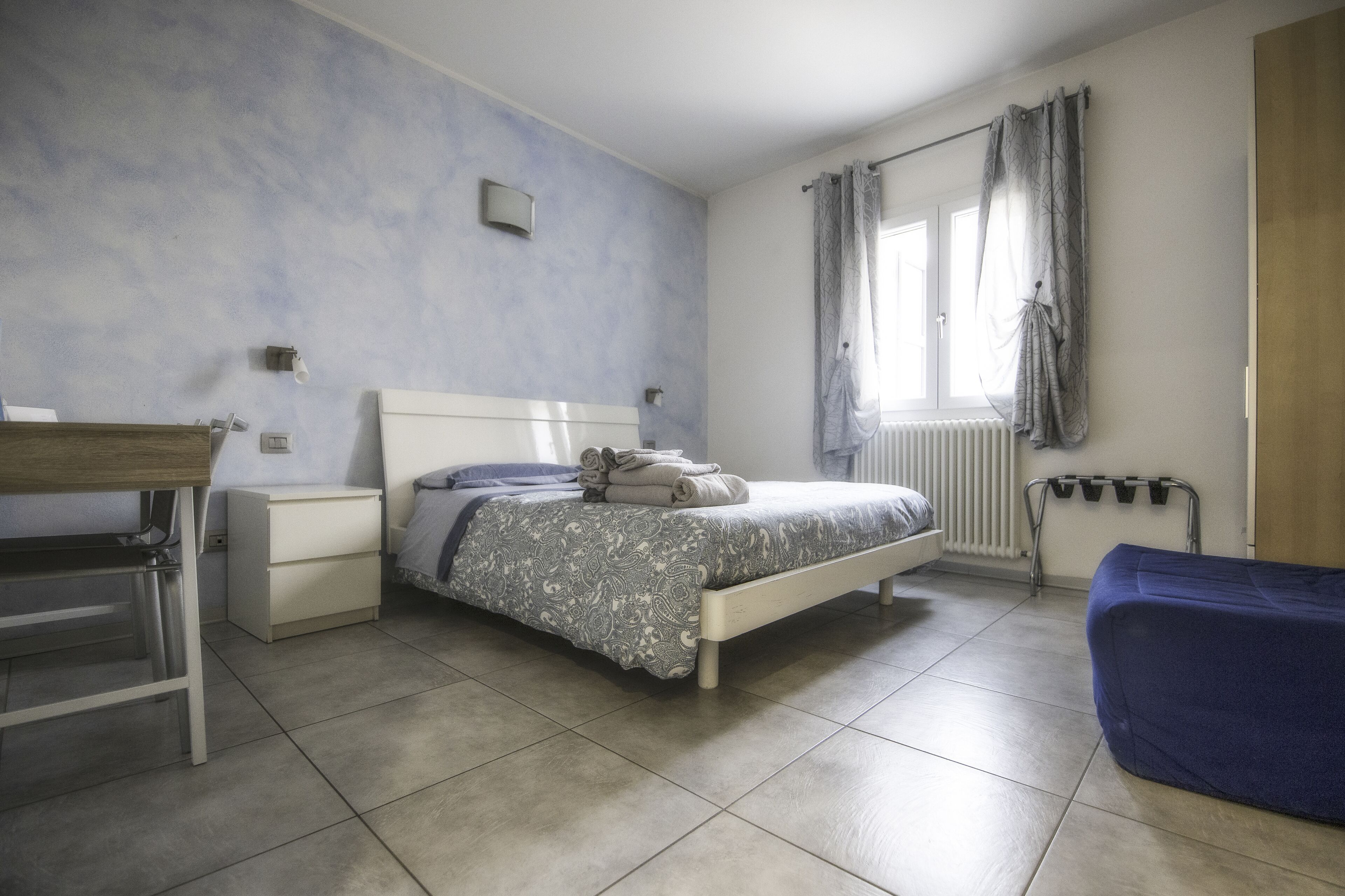 A bedroom with a bed, a desk, a chair, a radiator, and a window with curtains.