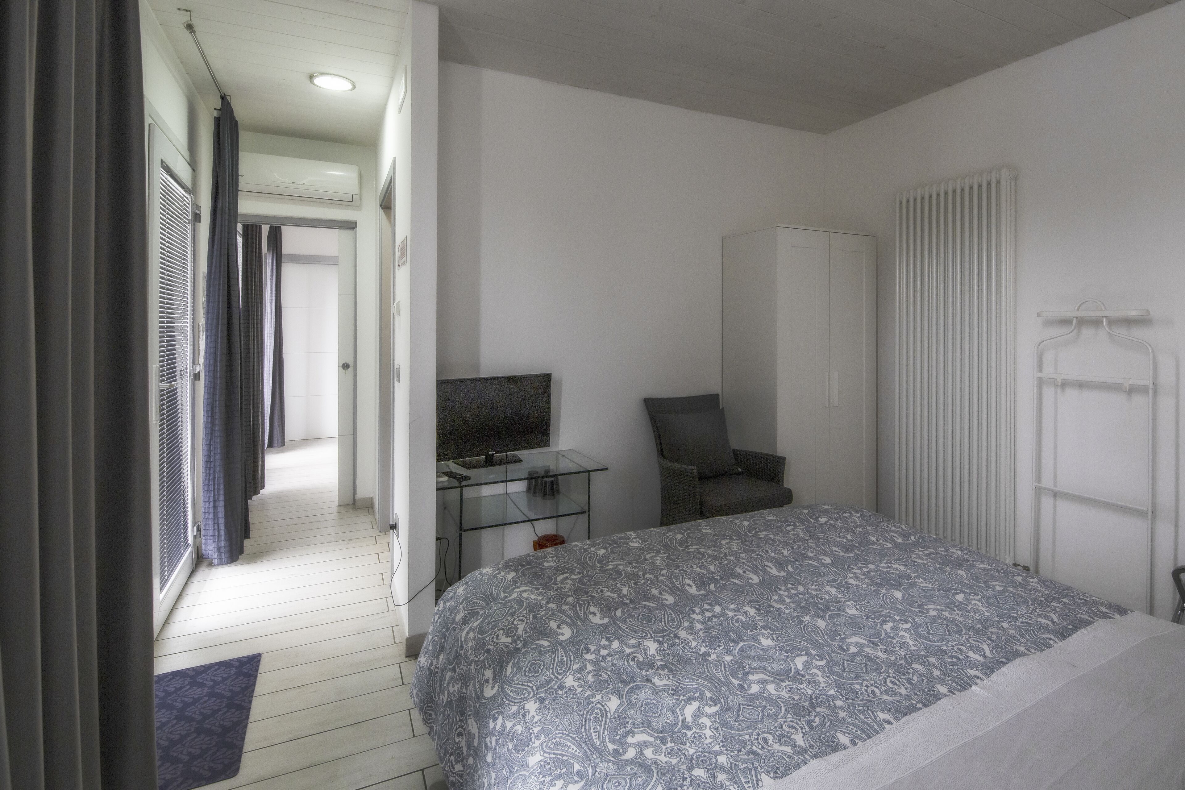 A bedroom with a bed, a television on a small table, a chair, a wardrobe, and a radiator.