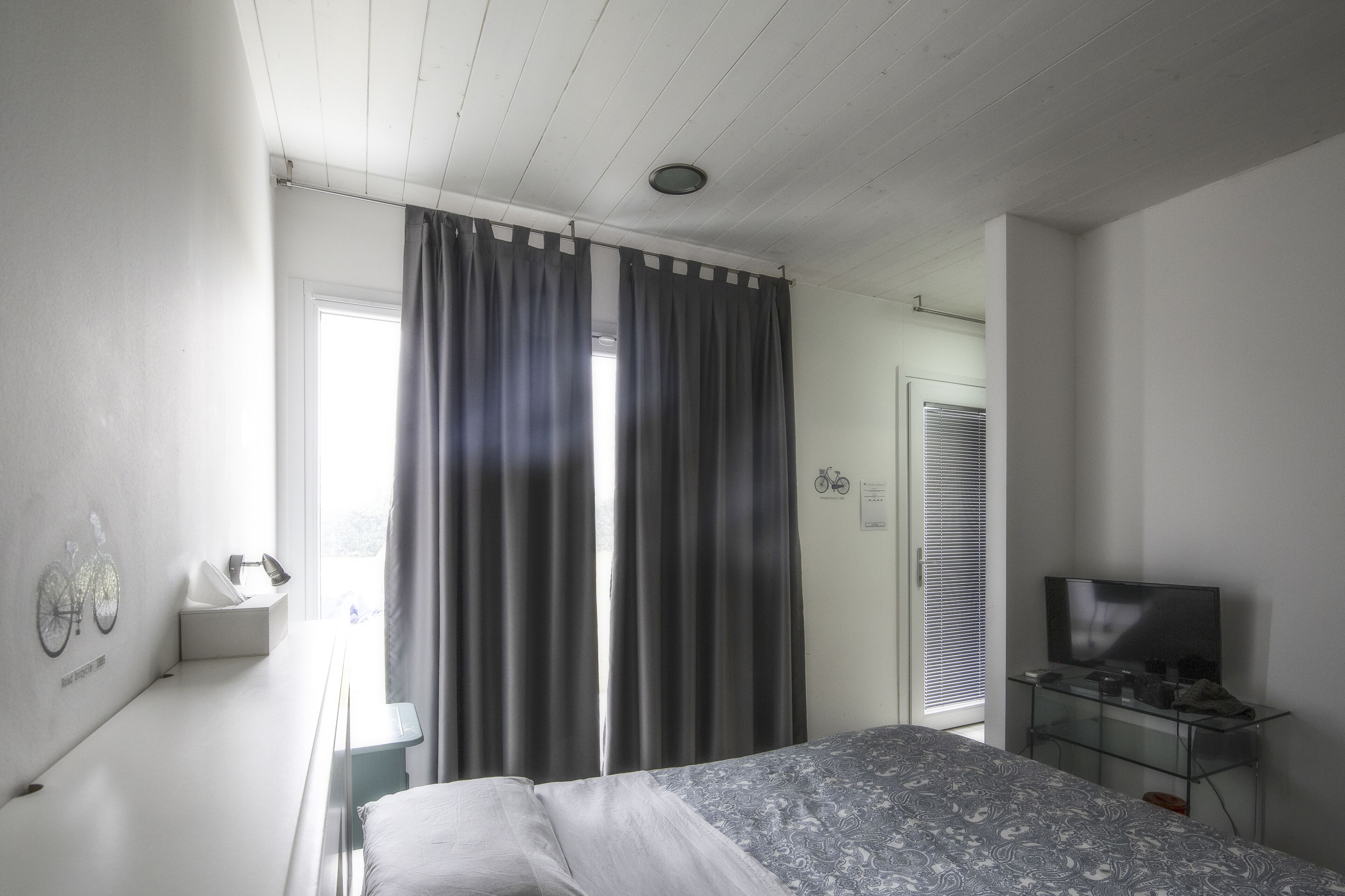 Une chambre avec un lit, un téléviseur posé sur un meuble, une fenêtre avec des rideaux, une décoration murale représentant un vélo, et une porte ornée d’un symbole de vélo.