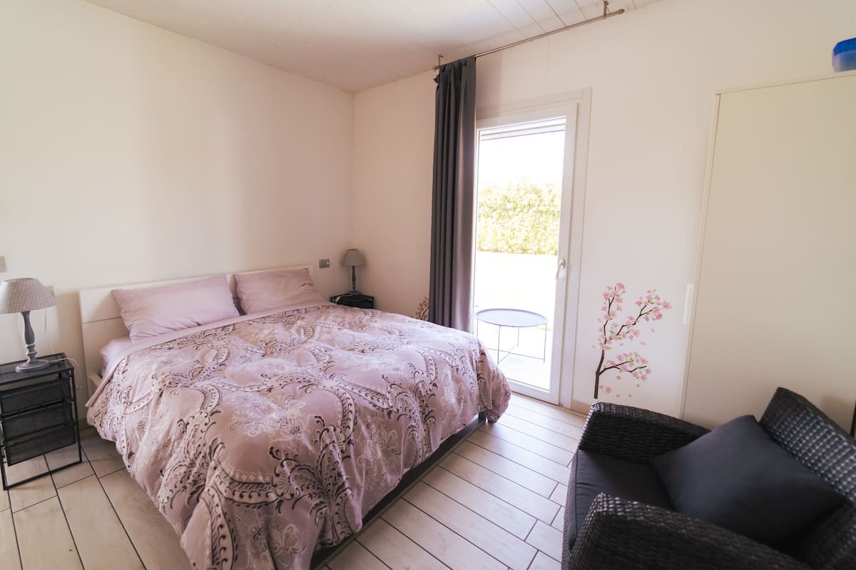A bedroom with a bed, a nightstand, a lamp, a chair, and a door to a balcony.