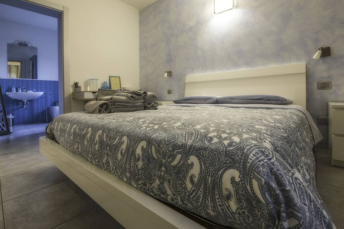 A modern bedroom with a large bed, a nightstand, a wall-mounted lamp, and a bathroom visible through an open door.