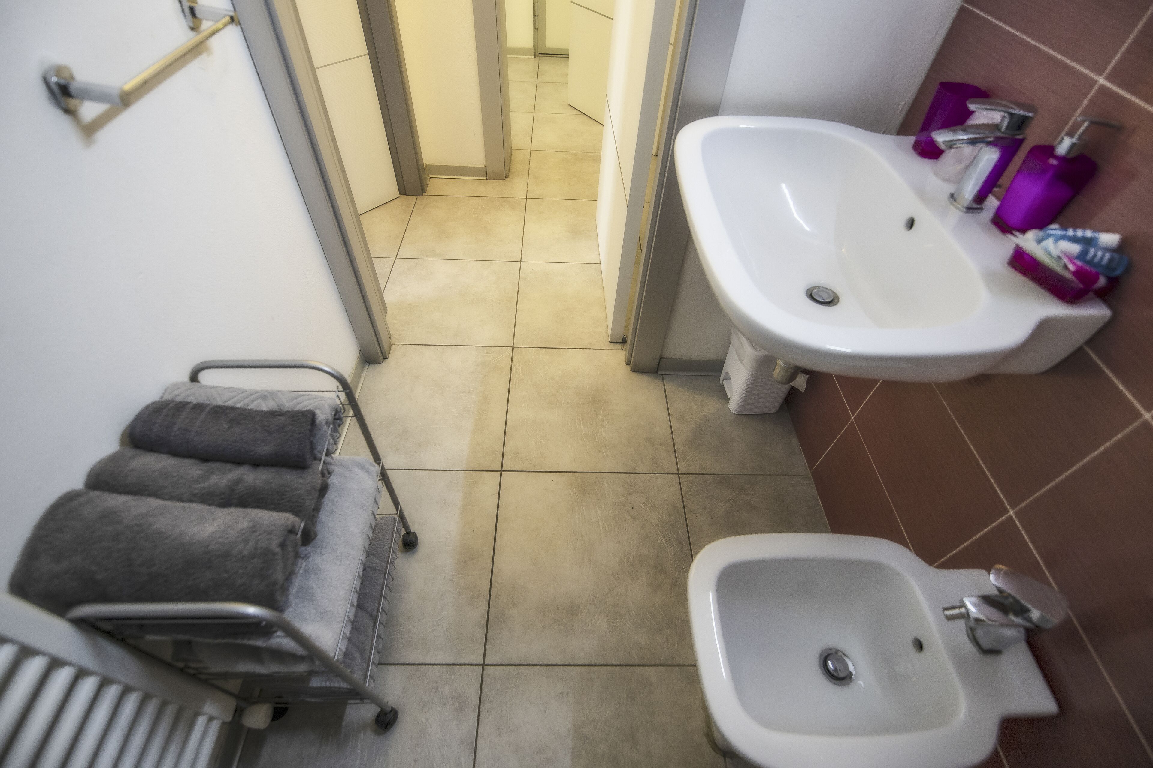 A bathroom with a white sink, a bidet, a towel rack, and a shower chair.