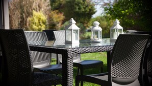 Una mesa con cubierta de vidrio, sillas de mimbre, faroles blancos y vistas a árboles y césped.