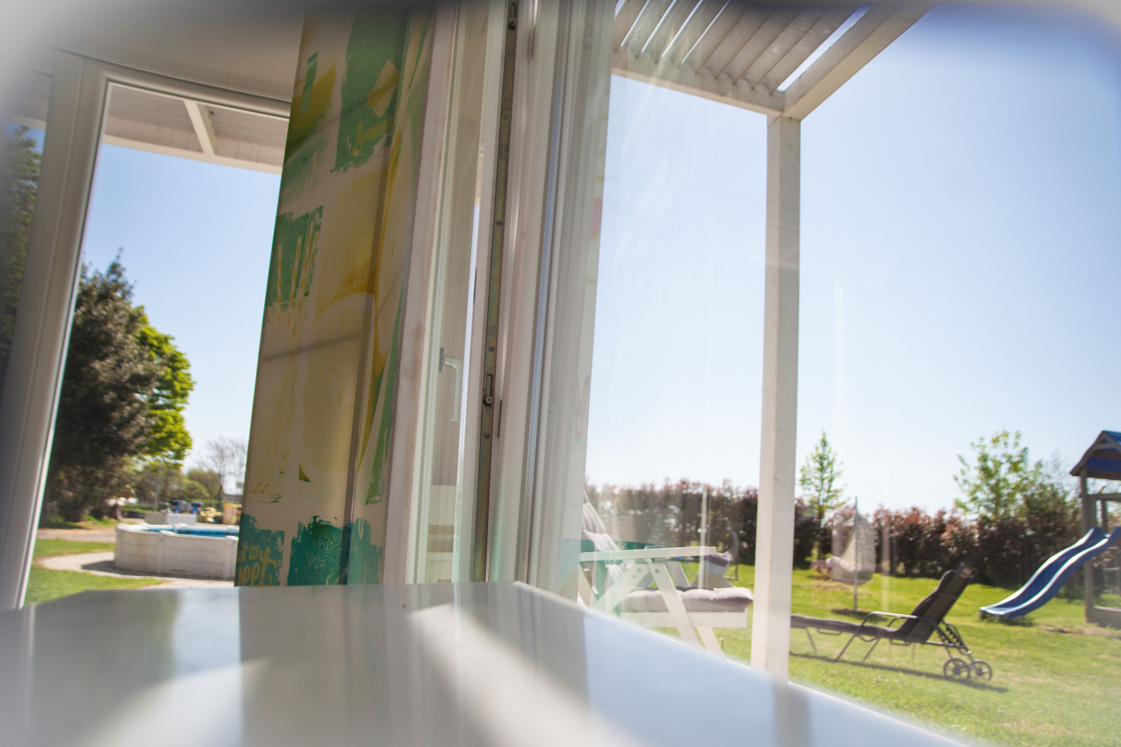 A view through a window with a clear sky, a grassy yard, and a playground with a slide.