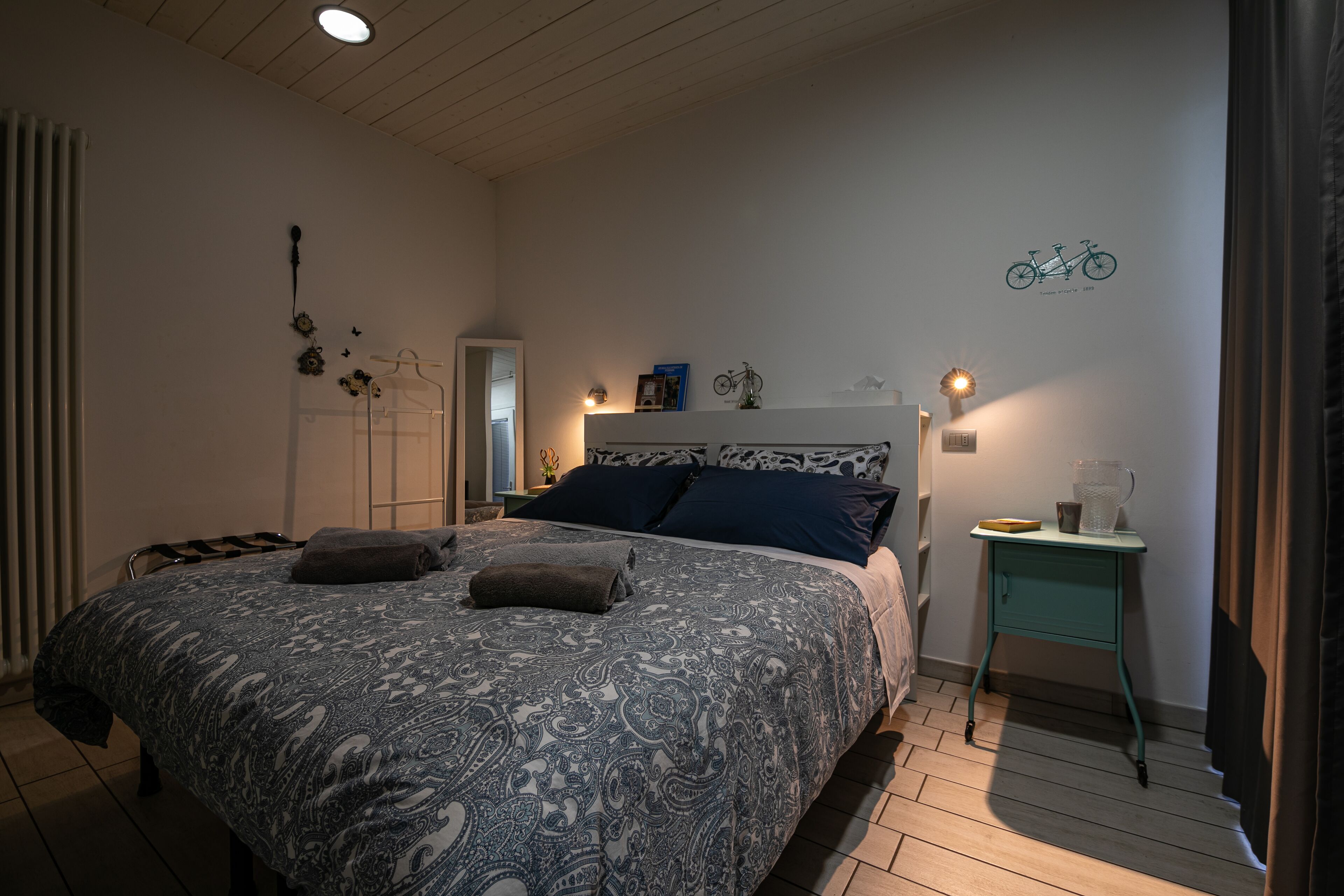 Une chambre à coucher avec une couvre-lit à motifs, une table de chevet avec une lampe et une illustration de vélo accrochée au mur.