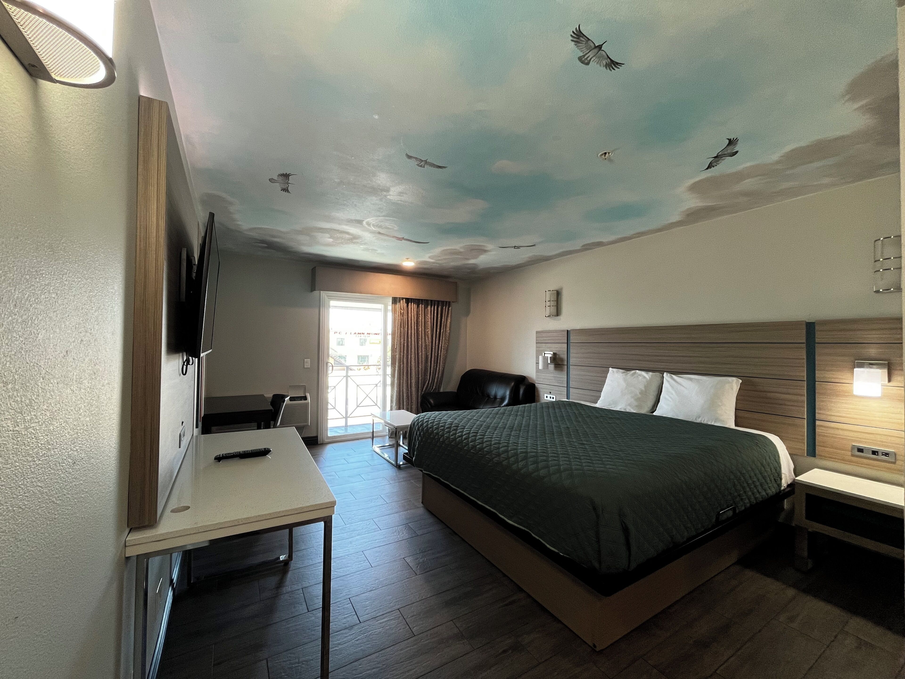 A hotel room with a bed, a desk, a TV, a chair, and a ceiling mural.