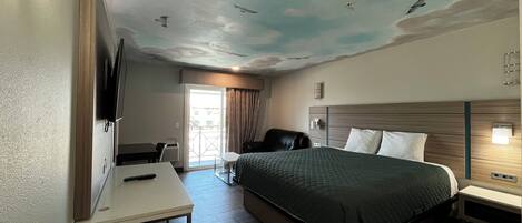 A hotel room with a bed, a desk, a TV, a chair, and a ceiling mural.