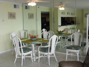 A dining area with a glass table and white chairs, a ceiling fan, and a kitchen visible through a glass partition.