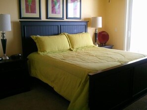 A neatly made bed with a blue headboard and two pillows, a bedside table with a lamp, and a framed picture on the wall. - Tern Key Realty & Rentals (Pensacola)