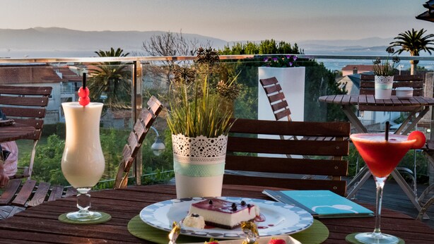 A wooden deck with a table set for a meal, featuring a dessert plate, a martini glass, and a tall drink. - Hotel & Apartamentos Cons da Garda (O Grove)