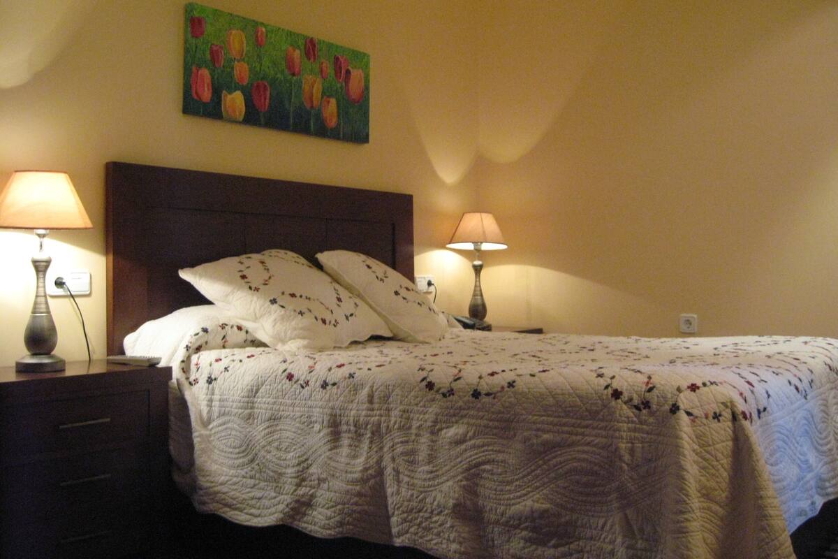 A neatly made bed with floral bedding, two bedside lamps, and a painting of tulips on the wall.