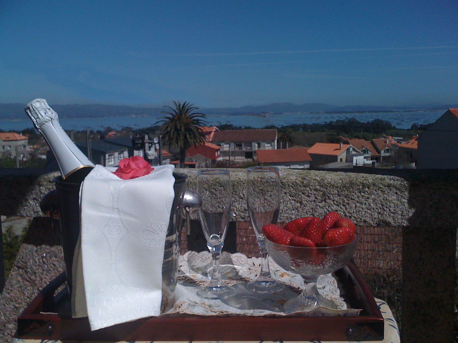 Una mesa con una botella de champán, una copa y un bol de fresas en un balcón con vistas a un pueblo costero.