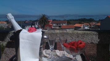 A table with a bottle of champagne, a glass, and a bowl of strawberries on a balcony overlooking a coastal town.