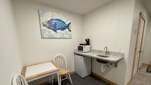 A compact kitchenette with a microwave, coffee maker, and sink. There is a small dining table with chairs and a painting of a fish on the wall. - The Inn at Old Town (Bandon)