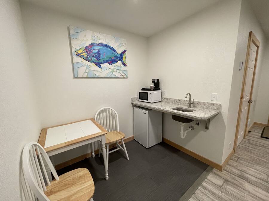 A compact kitchenette with a microwave, coffee maker, and sink. There is a small dining table with chairs and a painting of a fish on the wall.