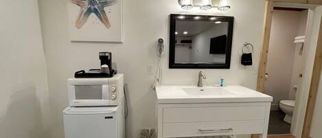 A modern bathroom with a white sink, a mirror, a coffee maker, and a trash can.