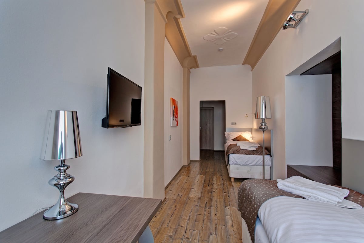 A hotel room with a wooden floor, a bed with white bedding, a bedside table with a lamp, a flat-screen TV mounted on the wall, and a door leading to another room.