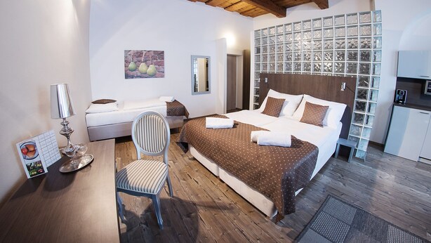 Habitación de hotel con techo de madera, una cama con colcha marrón estampada, una silla blanca, una mesita con lámpara y una zona de cocina con refrigerador y alacenas.