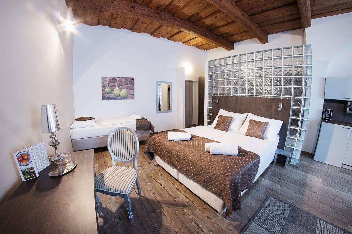 A hotel room with a wooden ceiling, a bed with a brown patterned bedspread, a white chair, a small table with a lamp, and a kitchen area with a refrigerator and cabinets.