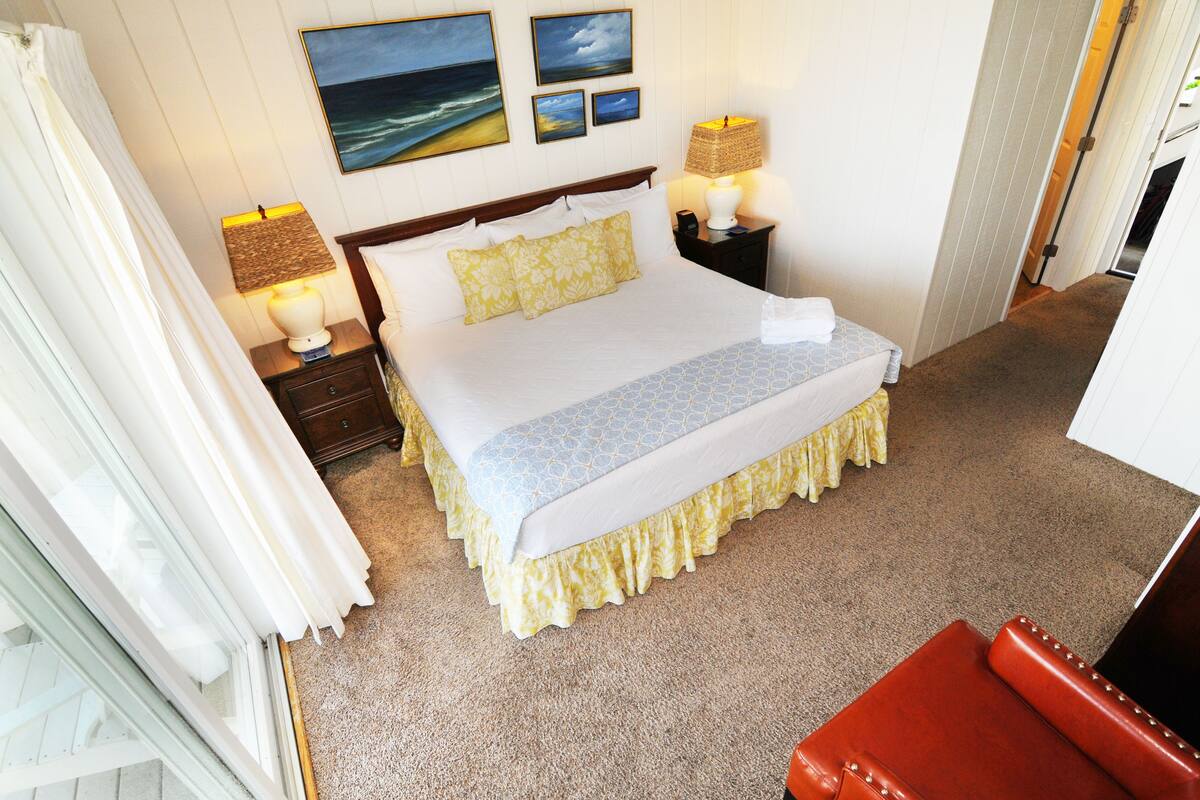 A hotel room with a bed, two bedside tables with lamps, a red leather chair, and a view of the ocean through the window.