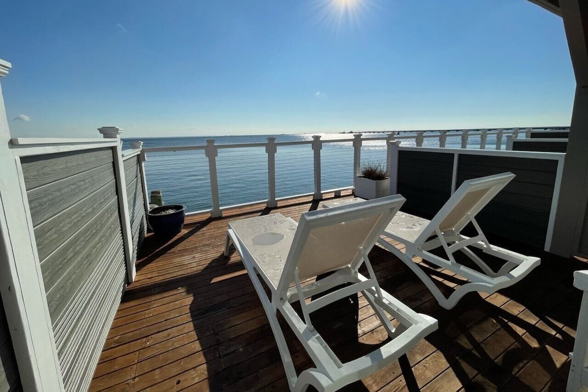A wooden deck with two white lounge chairs and a table, overlooking a body of water with a clear sky.