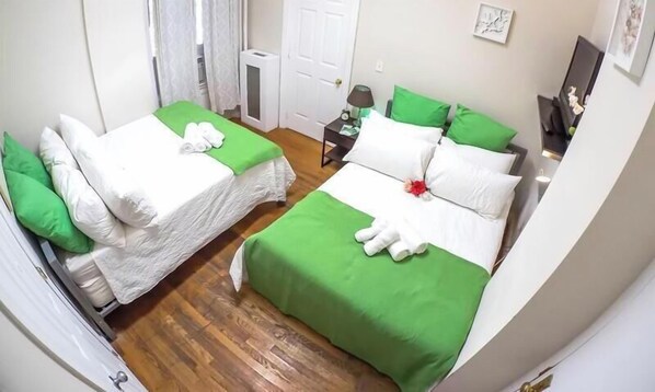 Una habitación de hotel con dos camas, ropa de cama verde y blanca, una mesita de noche con lámpara y una puerta.
