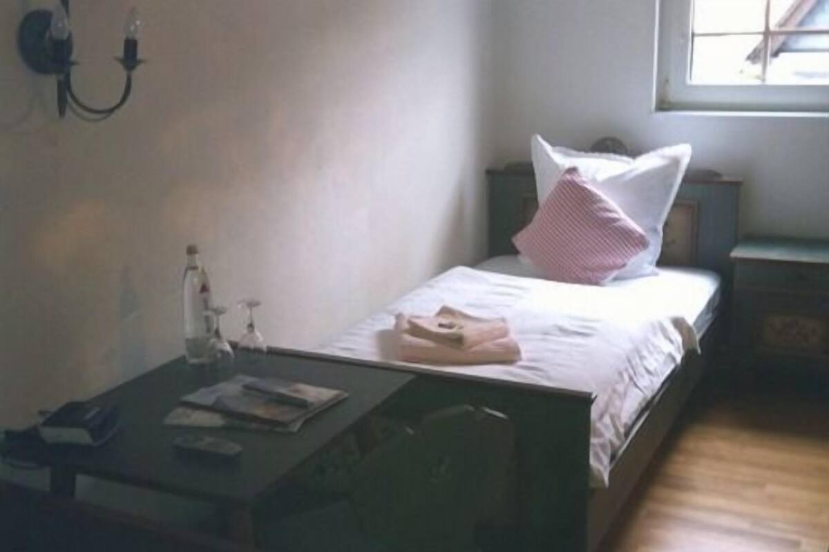 A small, simple room with a bed, a nightstand, a table with a bottle and a book, and a window.