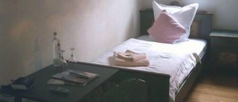 Una habitación pequeña y sencilla con cama, mesita de noche, una mesa con una botella y un libro, y una ventana.