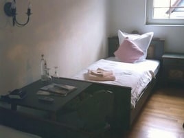Una habitación pequeña y sencilla con cama, mesita de noche, una mesa con una botella y un libro, y una ventana.