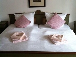 Una cama bien hecha con sábanas blancas, dos almohadas a cuadros rosas y dos toallas dobladas.