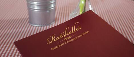 A dining table with a metal cup holding utensils, a vase with a red rose, and a menu titled "Ratsschäcker."