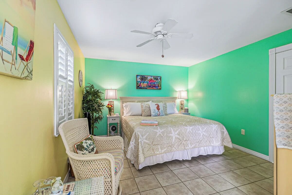 A bedroom with a bed, a chair, a nightstand, a ceiling fan, and a painting on the wall.