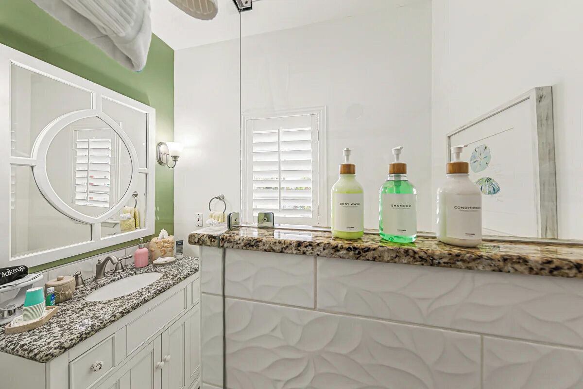 Une salle de bain avec un comptoir en granit, un meuble-lavabo blanc et un grand miroir. On y trouve trois flacons de produits de toilette posés sur le comptoir.