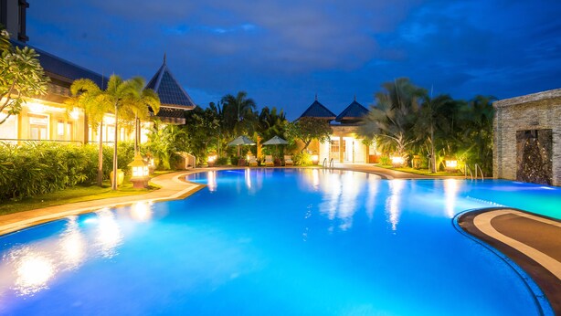 A swimming pool with a curved edge, surrounded by palm trees and a building with a peaked roof. - Hotel Shwe Pyi Thar (Mandalay)