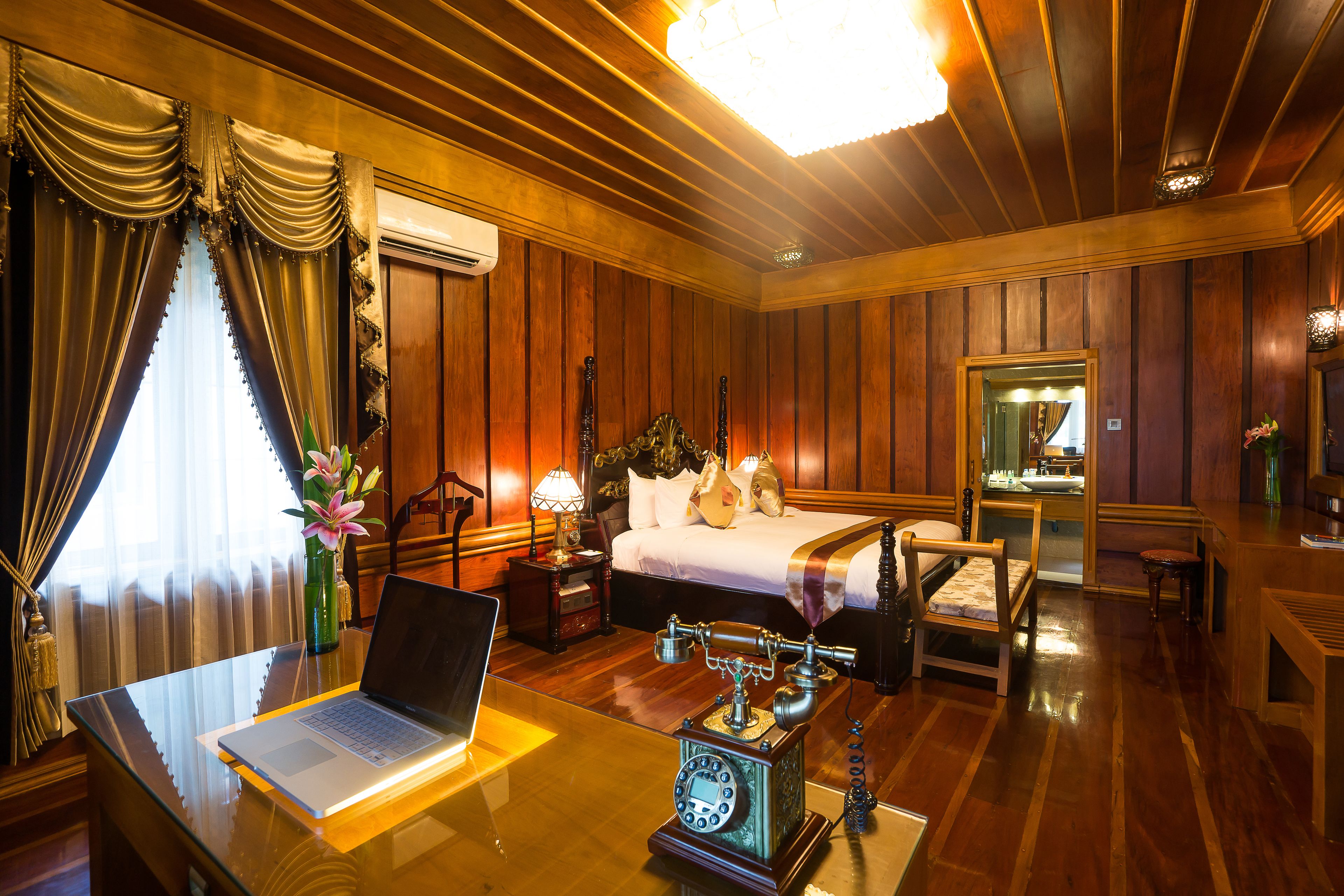 Una habitación de hotel con pared de paneles de madera, una cama con sábanas blancas, un escritorio con una computadora portátil, un teléfono antiguo y una araña.