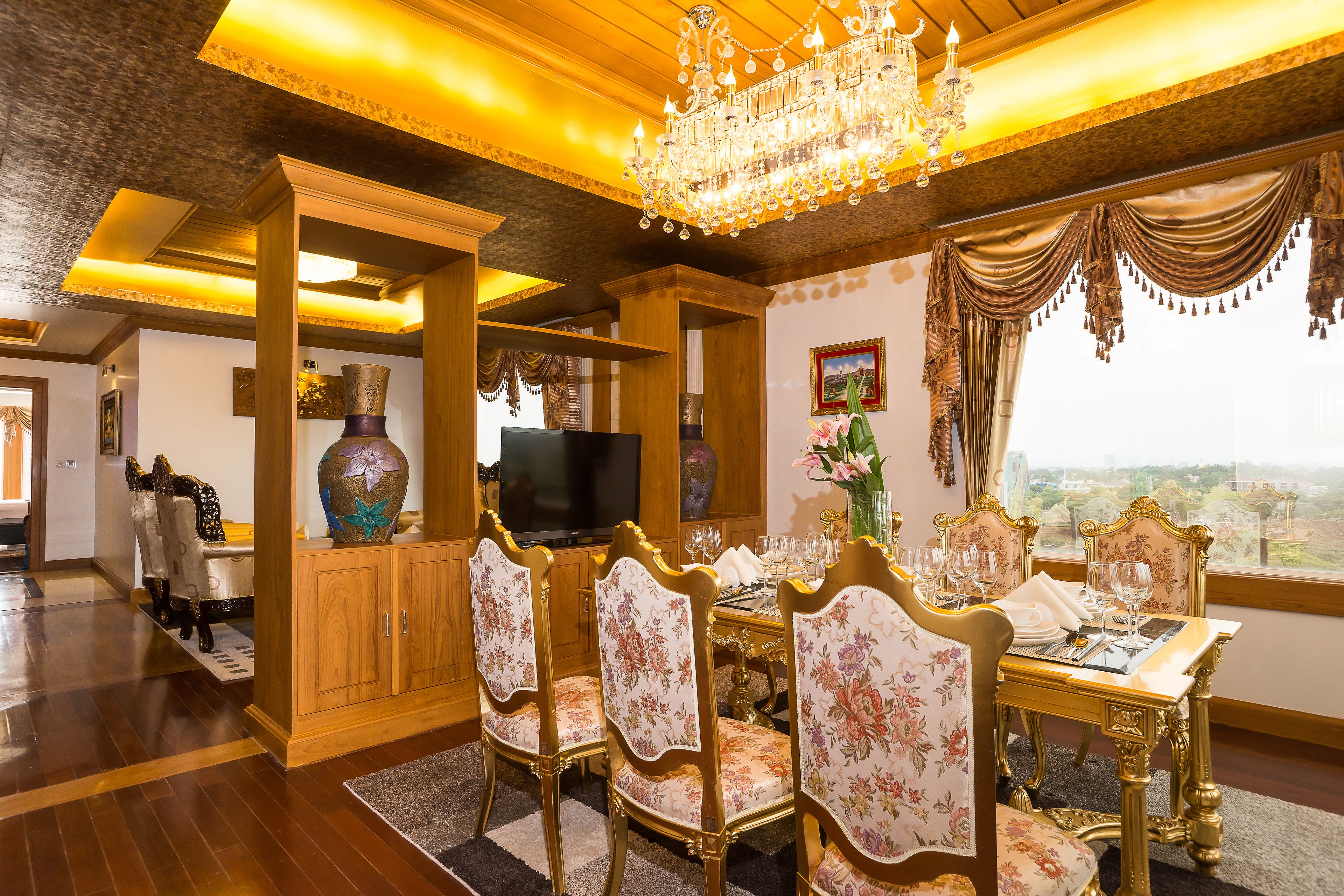A dining area with a chandelier, floral upholstered chairs, a wooden cabinet, and a television.