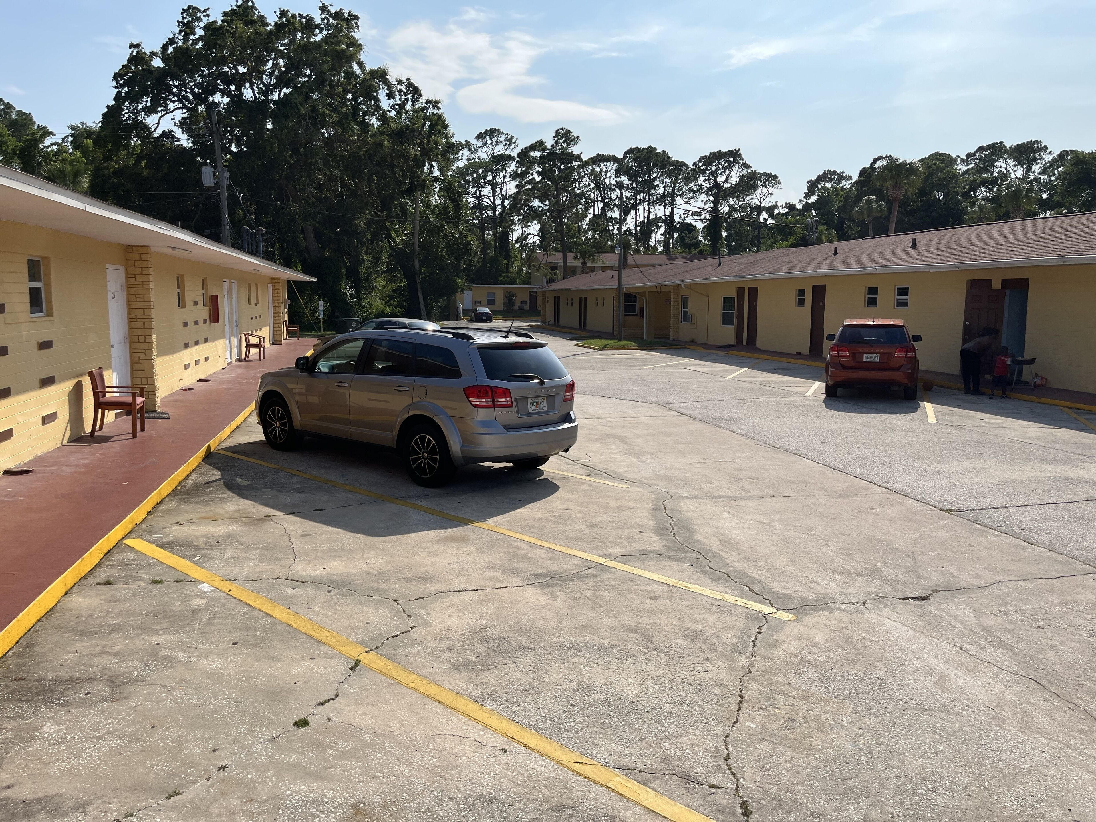 A parking lot with two cars, a row of motel-style buildings, and a red brick pathway.