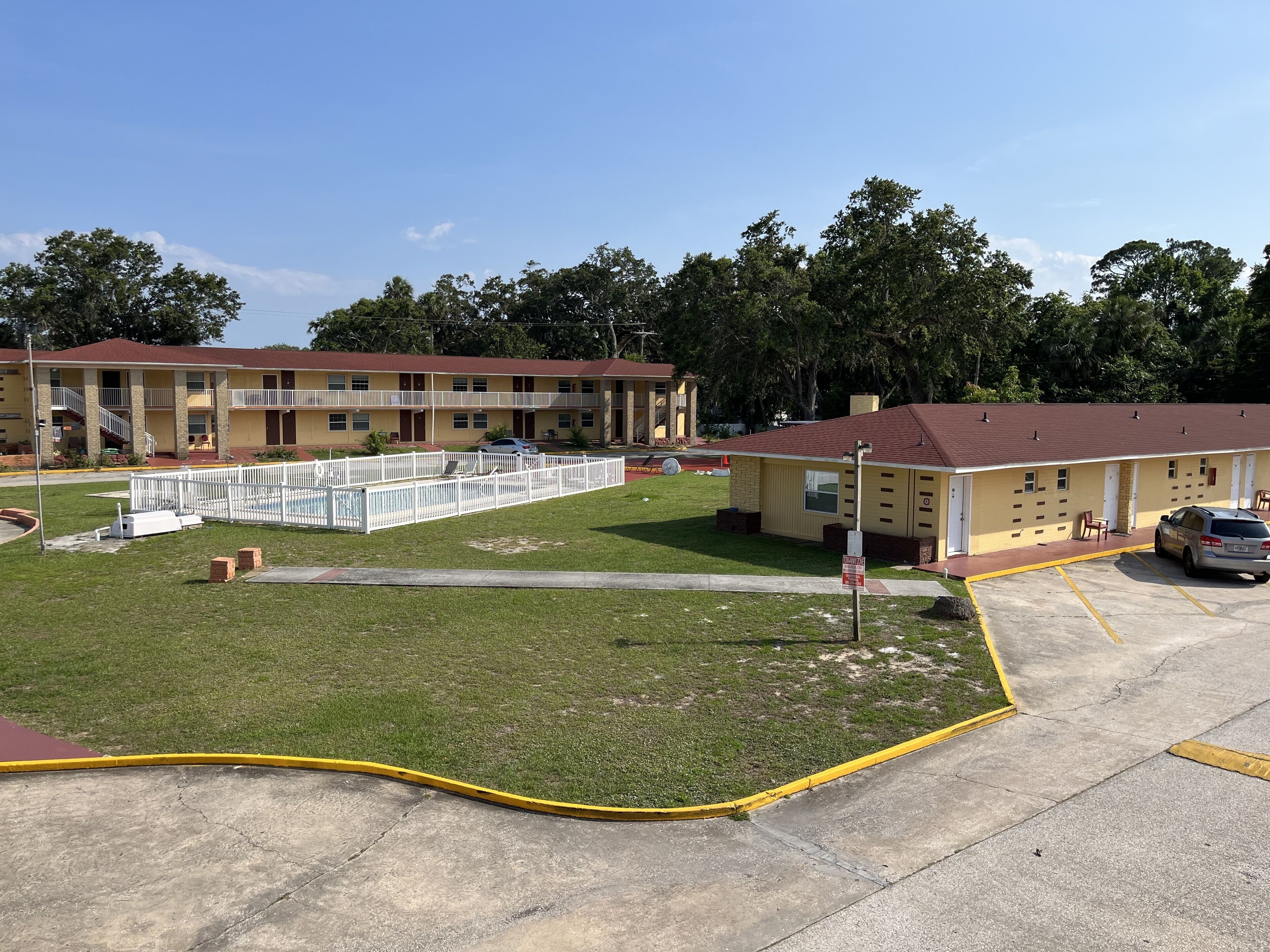 Um motel com área de piscina cercada, estacionamento e um pequeno edifício adjacente à estrutura principal.