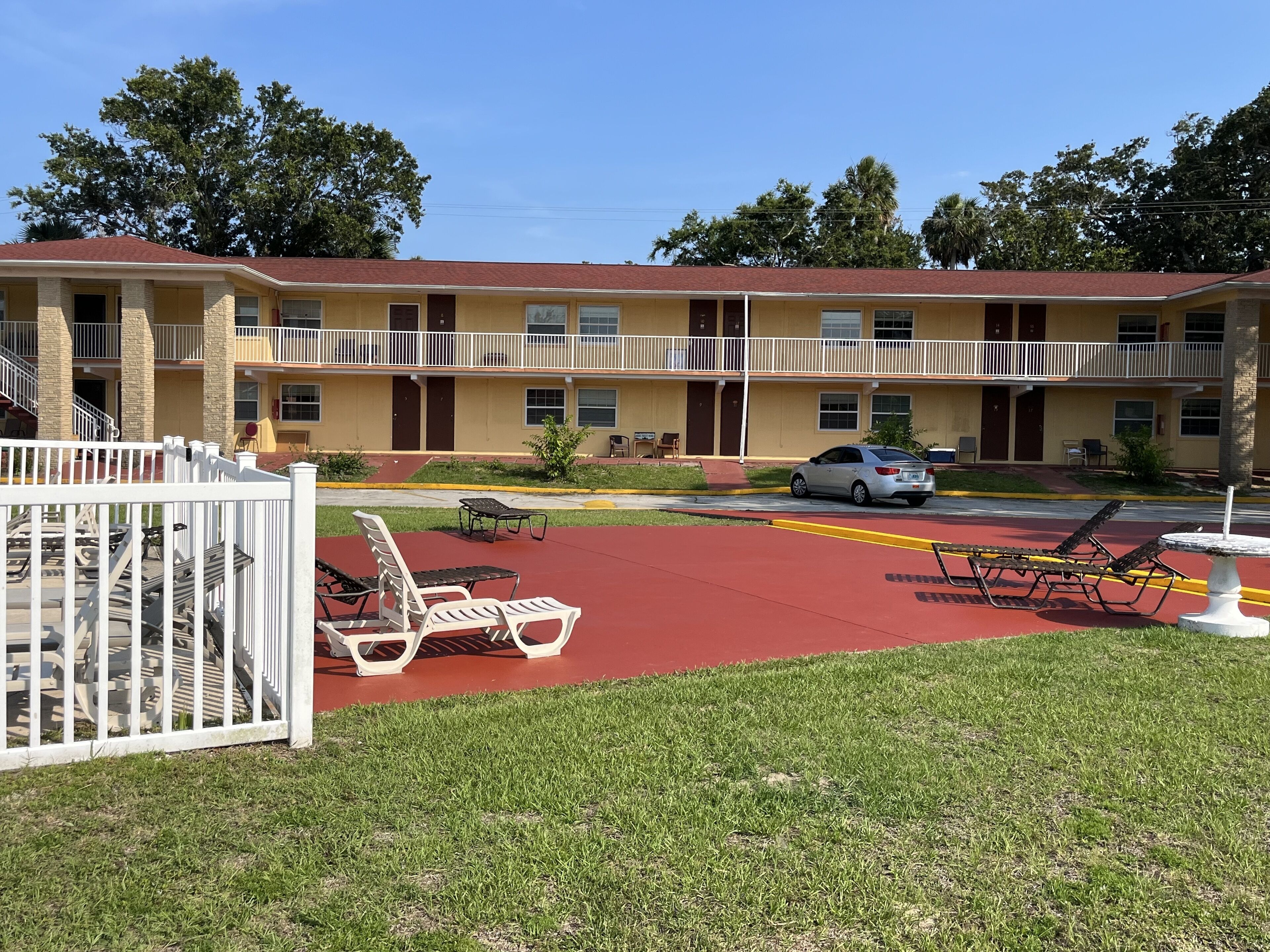 A motel with a red parking area, outdoor seating, and a two-story building with balconies.
