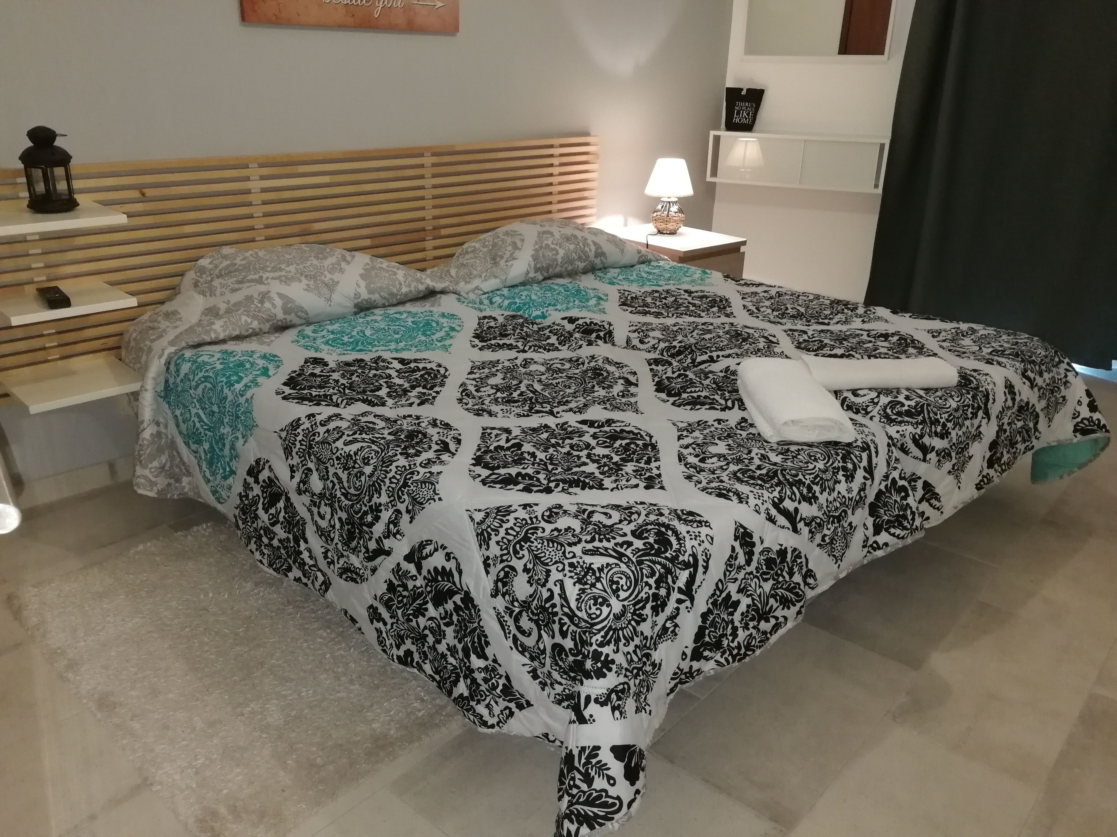 Un lit bien fait, recouvert d’une couvre-lit à motifs, une table de chevet avec une lampe et une étagère fixée au mur.