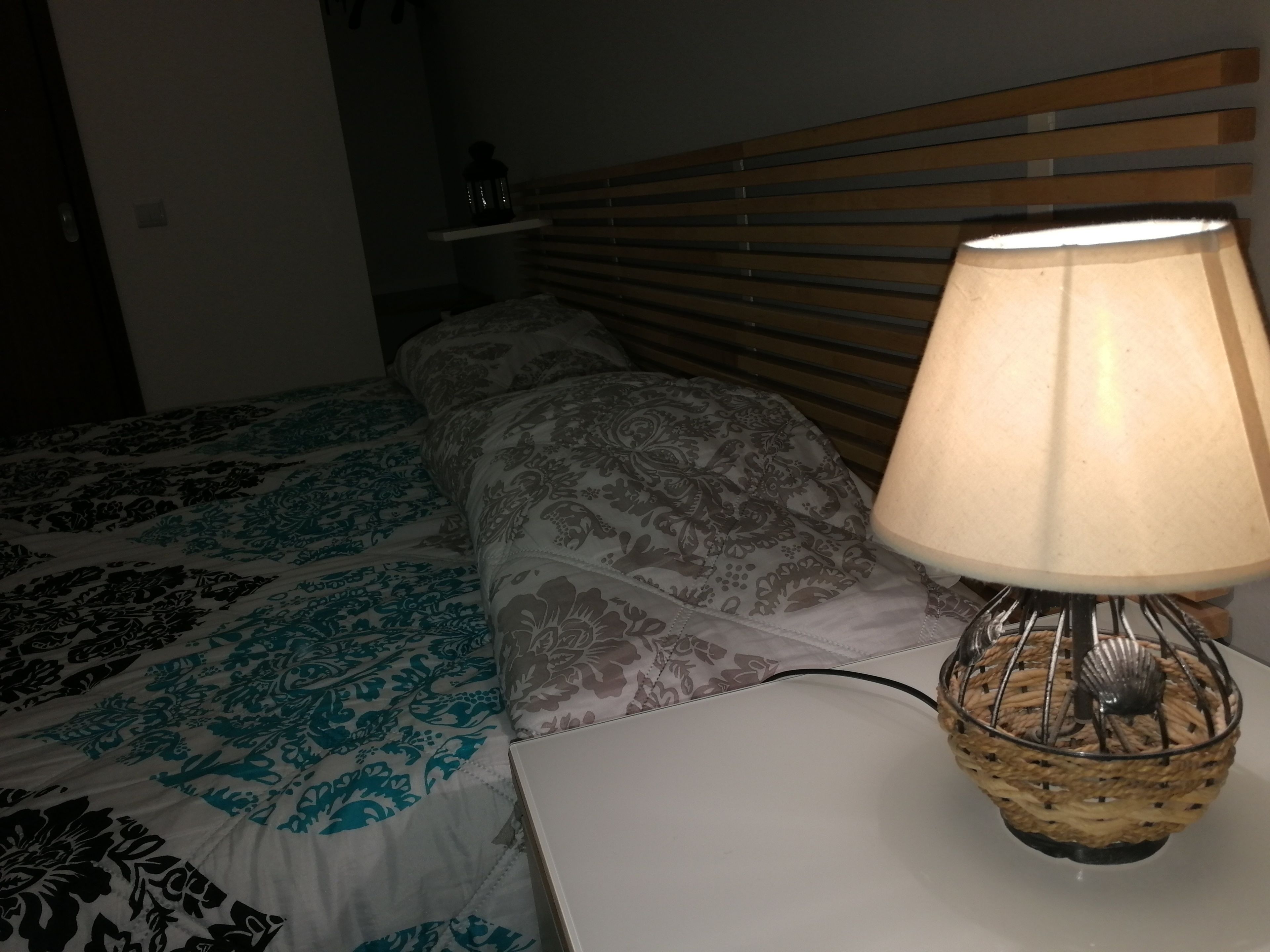 Un llit amb roba de llit estampada, una taula de nit amb una làmpada, i un panell de fusta al capçal.