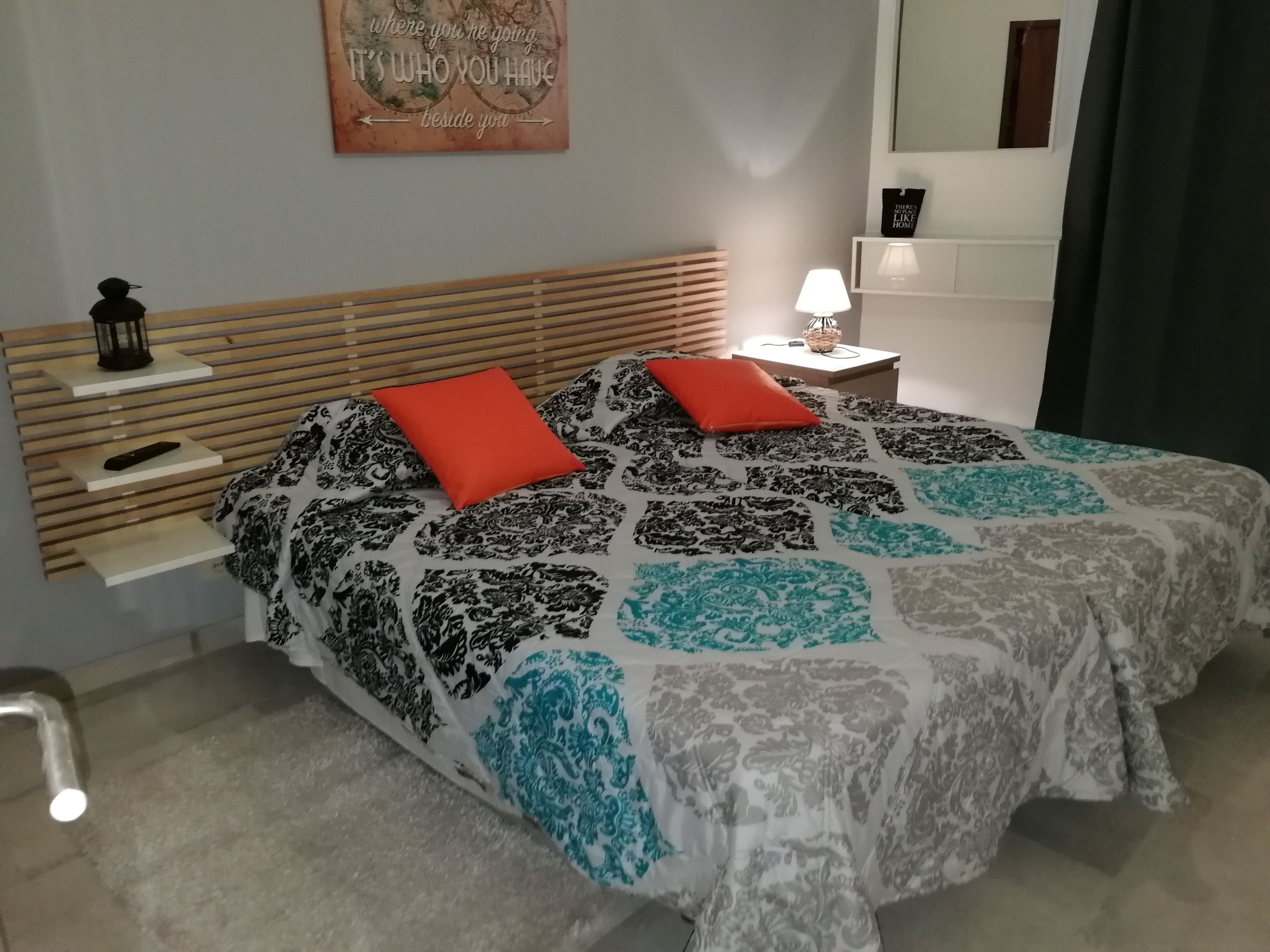 Ein Schlafzimmer mit einem Bett, zwei roten Kissen, einem Nachttisch mit Lampe, einer Wanduhr und einem motivierenden Zitat.