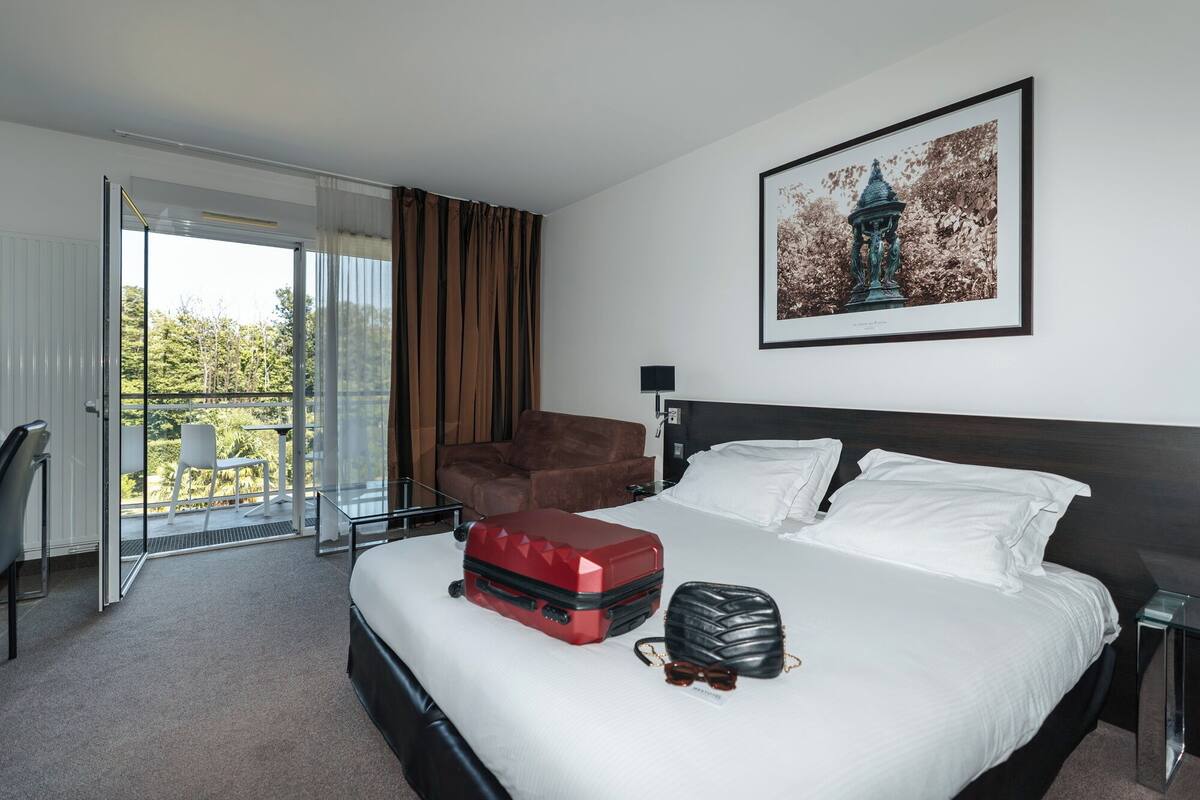 Habitación de hotel con una cama, una maleta roja, un bolso negro y un par de gafas de sol sobre la cama. Hay una silla marrón y una mesa de vidrio con una silla. Se ve un balcón con una puerta de vidrio y cortinas.