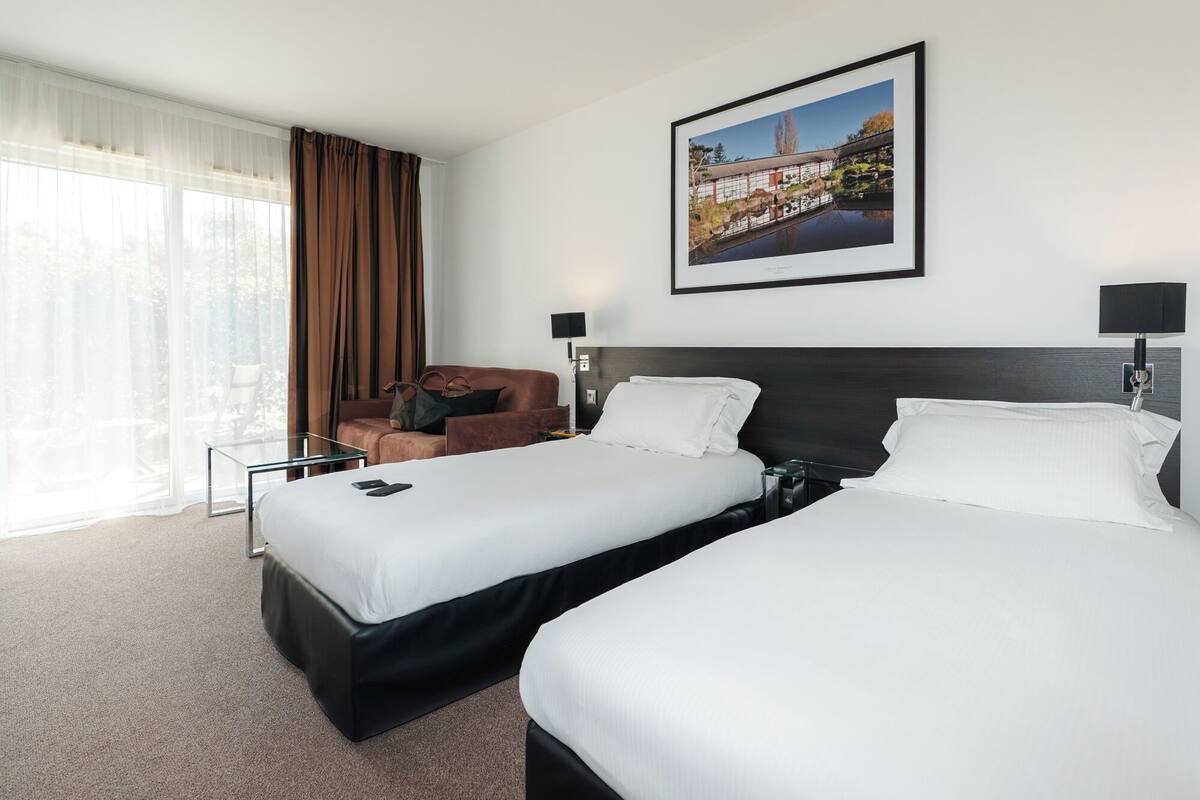 Una habitación de hotel con dos camas, una silla, un escritorio y un ventanal con cortinas.