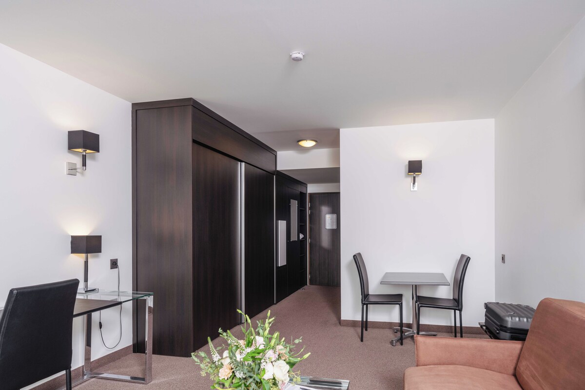 Une chambre d’hôtel moderne équipée d’un bureau, d’une chaise, d’un canapé, d’une table à manger avec des chaises et d’un vase à fleurs.