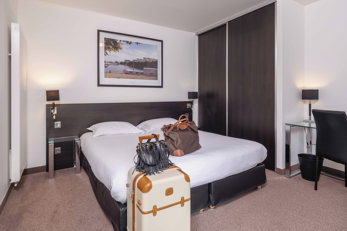 Habitación de hotel con una cama grande, una mesita de noche, una silla, un escritorio y un cuadro enmarcado en la pared.