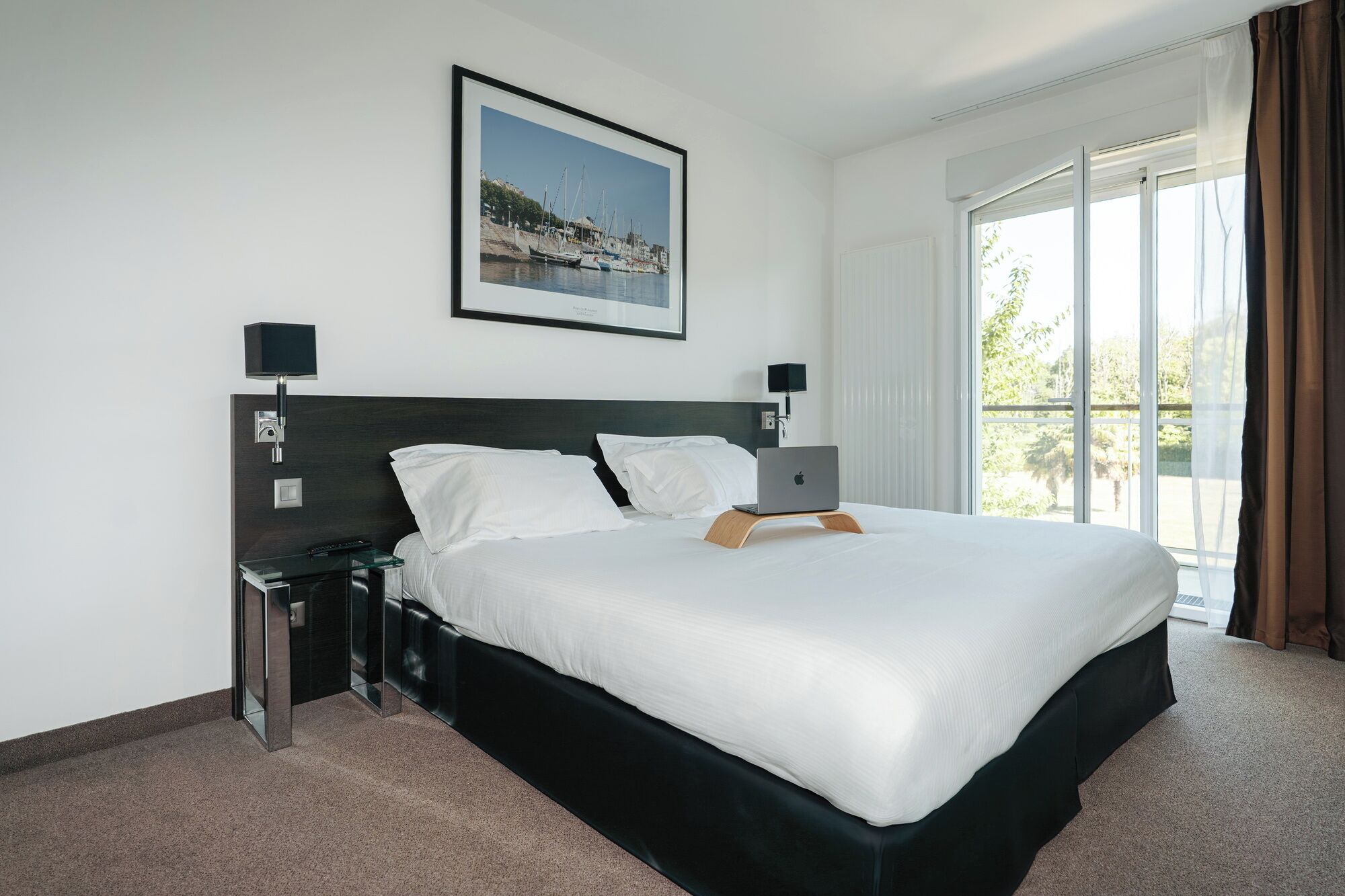 A hotel room with a large bed, a framed picture on the wall, a desk with a lamp, and a window with curtains.