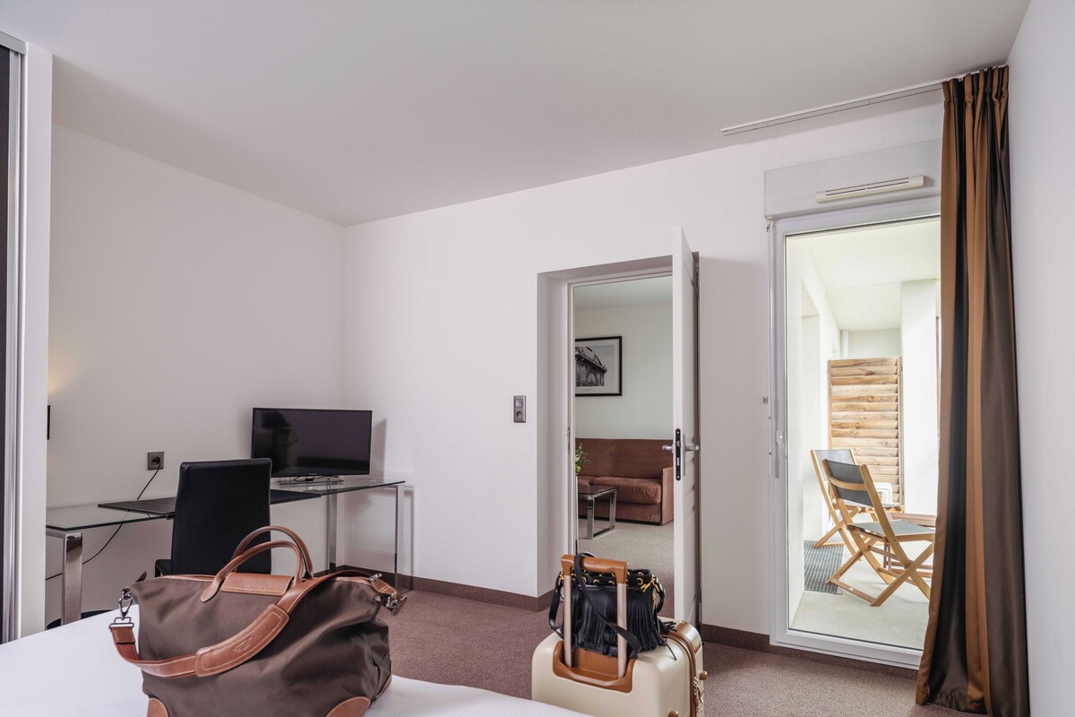 Une chambre d’hôtel avec un lit, un bureau équipé d’un ordinateur, une chaise, une valise et un sac à main.