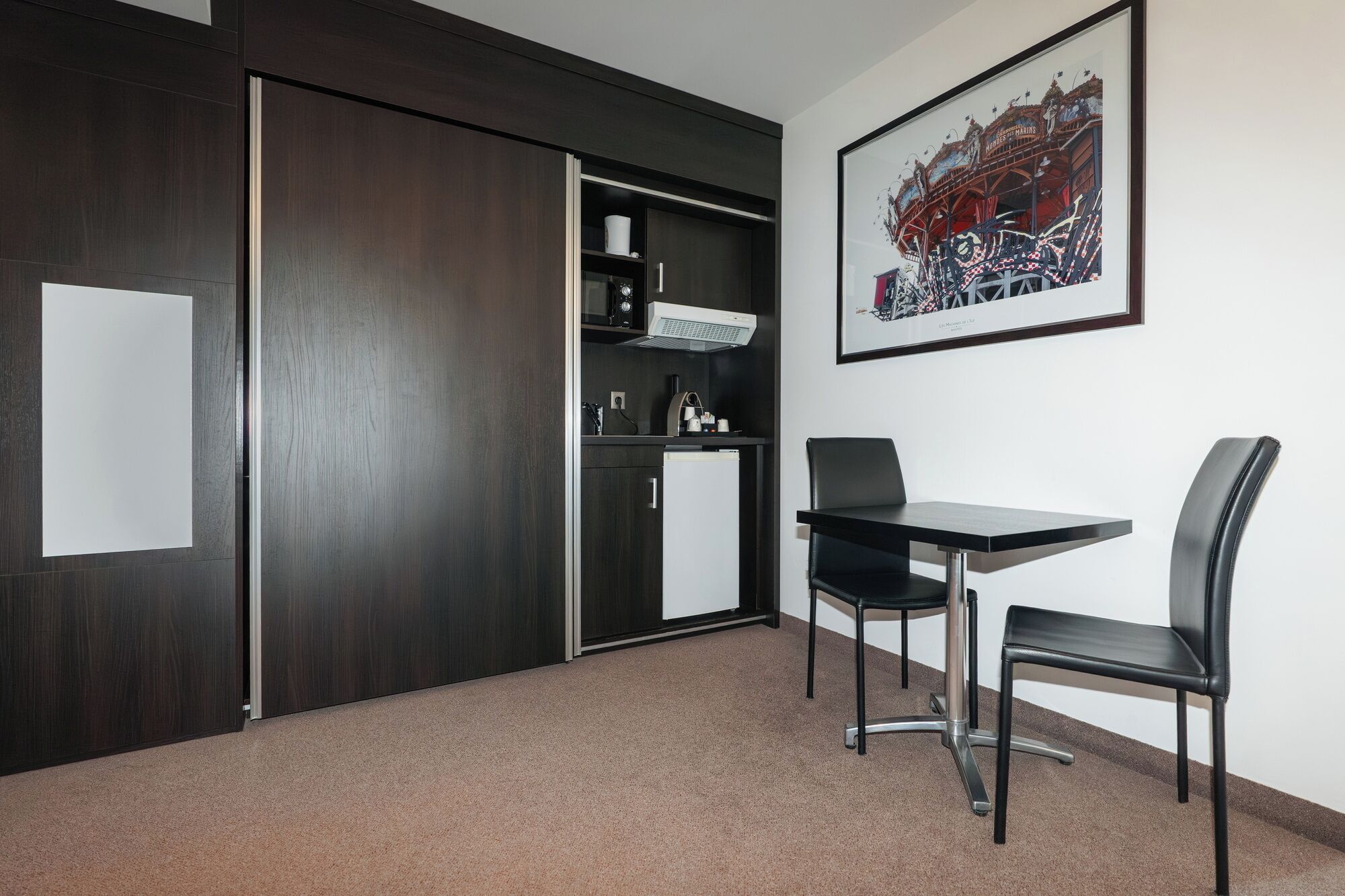 Ein Zimmer mit einem dunklen Holzschrank, einer kleinen Küchenzeile, einem gerahmten Bild an der Wand und einem Tisch mit zwei Stühlen.