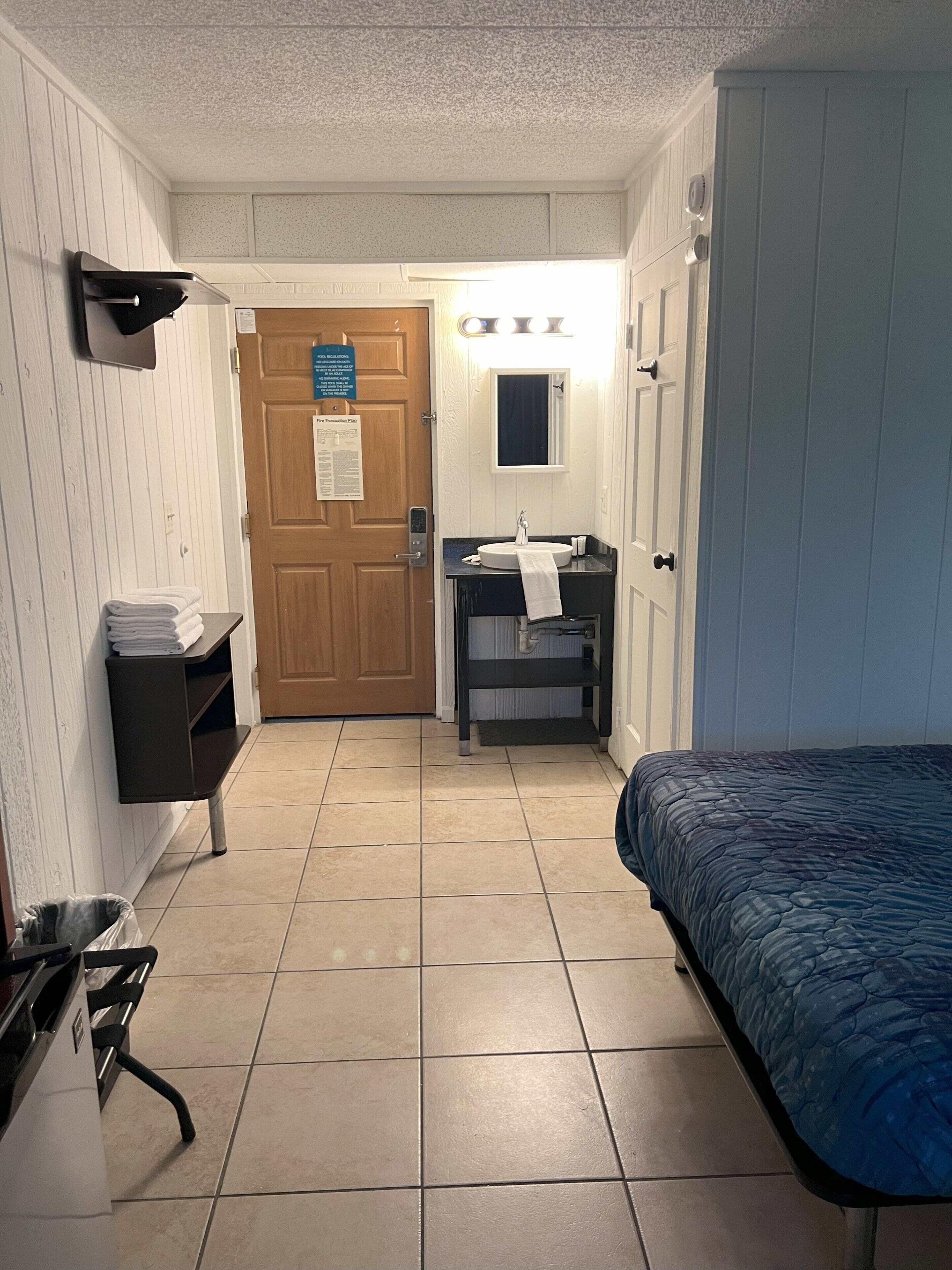 A hotel room with a bed, a desk with a sink, a mirror, and a door with a sign.